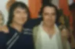 The Jim Twins smiling with arms around each other in a room. One wears a navy shirt with white stripes, the other in a beige jacket. Orange drapes behind.