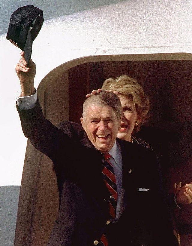 Ronald Reagan smiling, waving hat, with Nancy behind exiting plane.