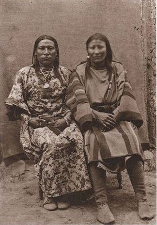 Two individuals in traditional attire, seated side by side against a plain backdrop. One wears a floral pattern, the other a striped design.