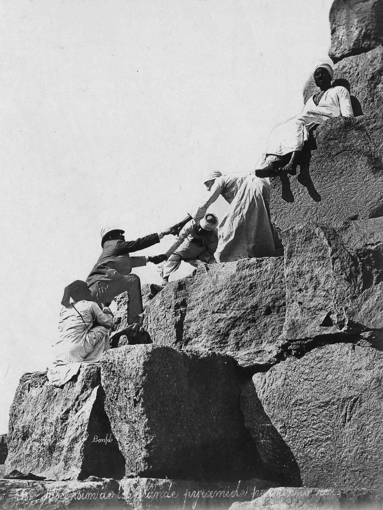 People helping each other climb a rocky cliff in a historical photo.