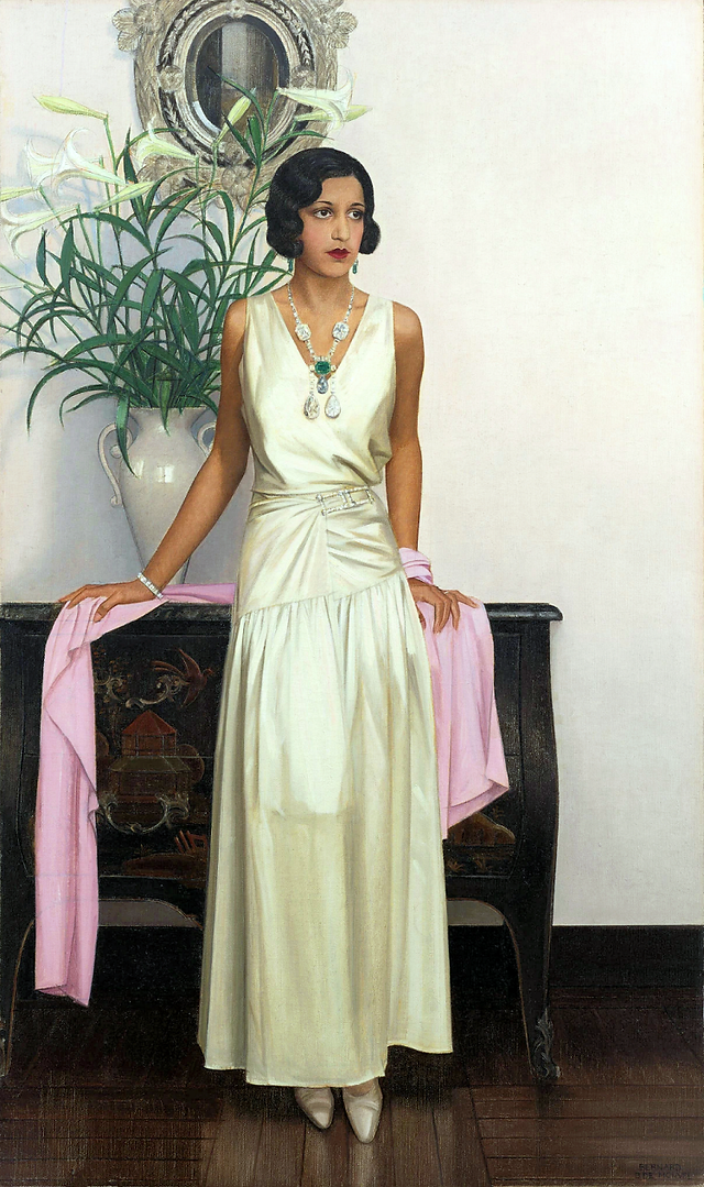 Woman in cream silk dress, wearing green pendant necklace, by ornate cabinet.