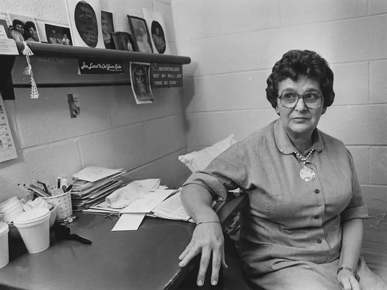 Older woman with glasses sits in an office by a cluttered desk. Black-and-white photos and motivational quotes decorate the wall. Mood is serious.