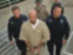 A handcuffed man in a beige prison uniform is escorted by three officers wearing dark uniforms. They walk along a concrete path outside.