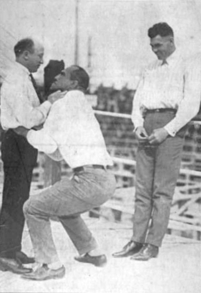 Three men outdoors; one man supports another in a squat position while a third man smiles nearby. All wear white shirts and dark pants.