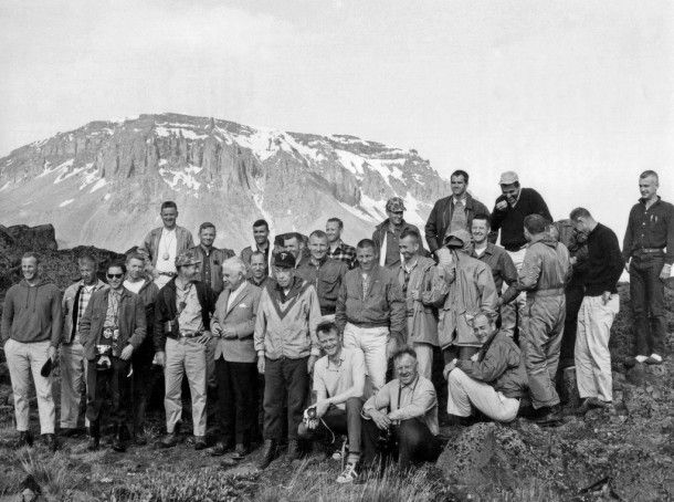 A group of men in jackets pose on rocky terrain with a snow-topped mountain in the background. The mood is cheerful and relaxed.