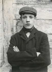 Serious young man in flat cap, jacket, arms crossed against wood.