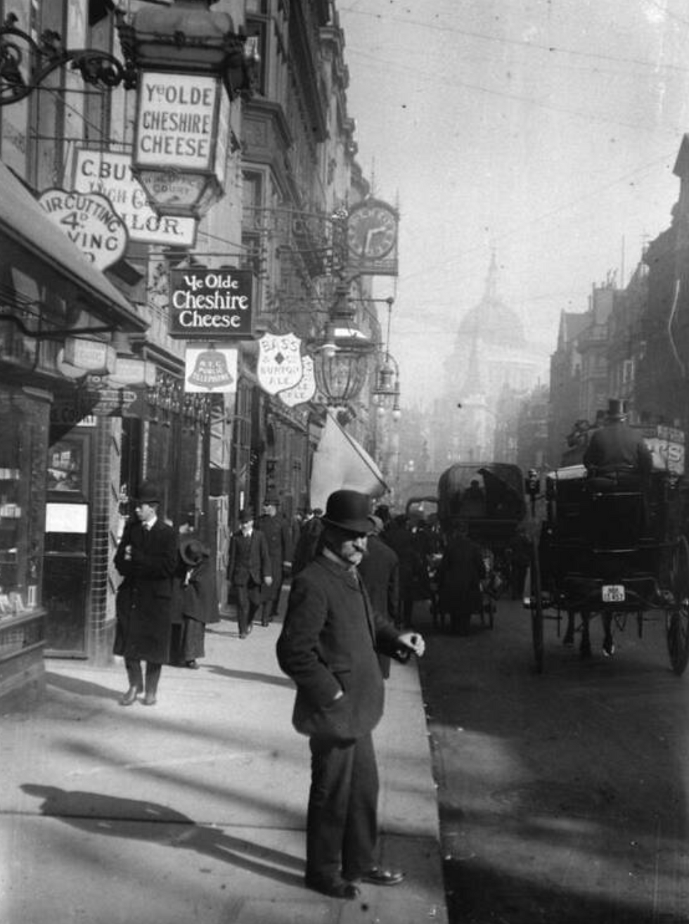 Historic street scene with people in coats and bowler hats, horse-drawn carriages, and shops. Sign reads "Ye Olde Cheshire Cheese." Vintage vibe.
