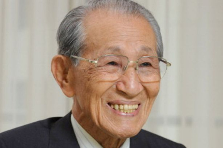 Elderly Asian man smiling, wearing glasses and a dark suit.