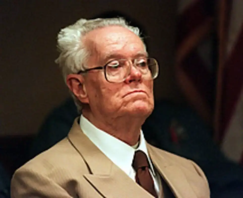 Elderly man with glasses in a tan suit sits with a stern expression against a dark background with a hint of a flag, conveying seriousness.