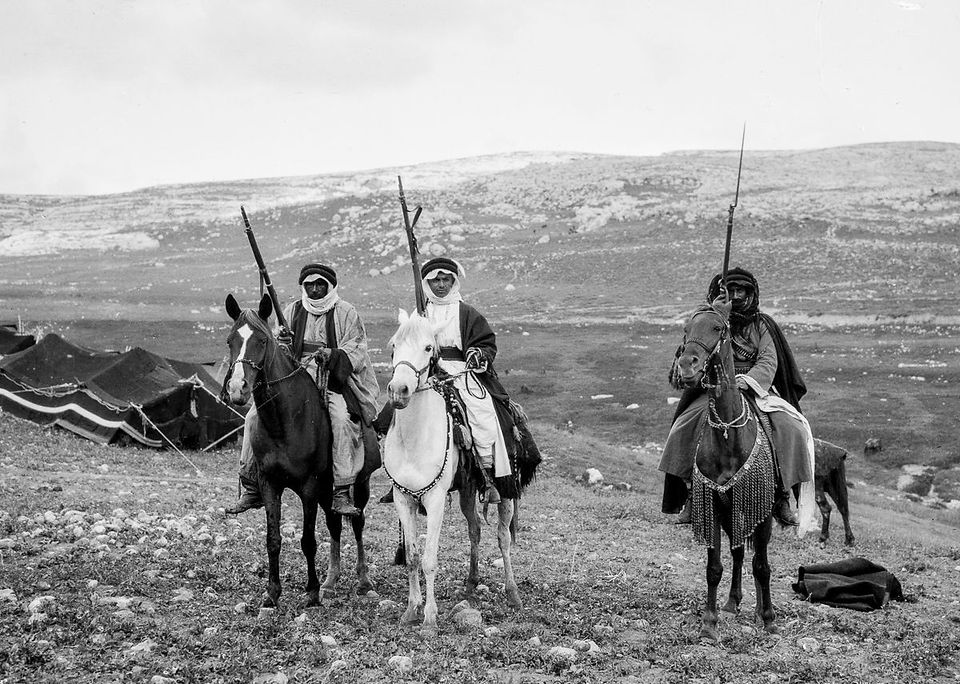 Black-and-white photograph of three Bedouin men on horseback, each holding a long rifle upright. They wear traditional robes and keffiyehs, with one mounted on a white horse and the others on dark horses. Behind them are tents and a rocky hillside. Captured in the early 20th century by the American Colony Photo Department.