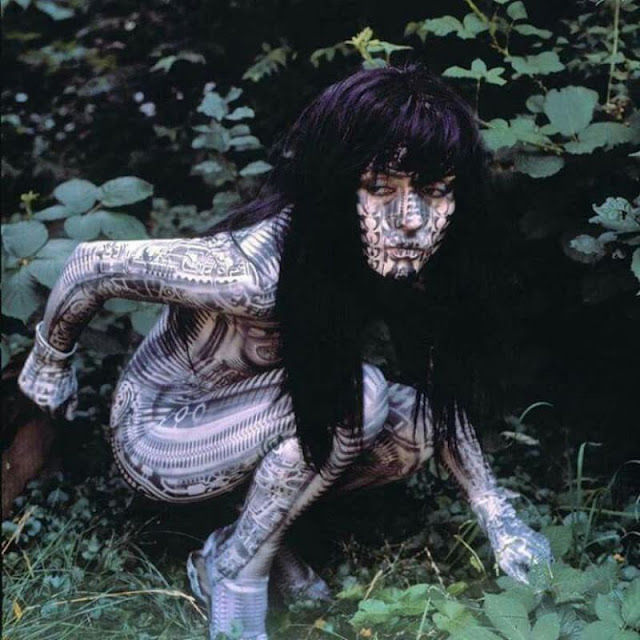 Person with intricate patterned body paint crouches in dense green foliage.