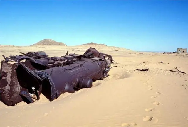 Rusty, overturned train engine in a sandy desert under a clear blue sky. Footprints lead to the horizon. Desolate, abandoned scene.