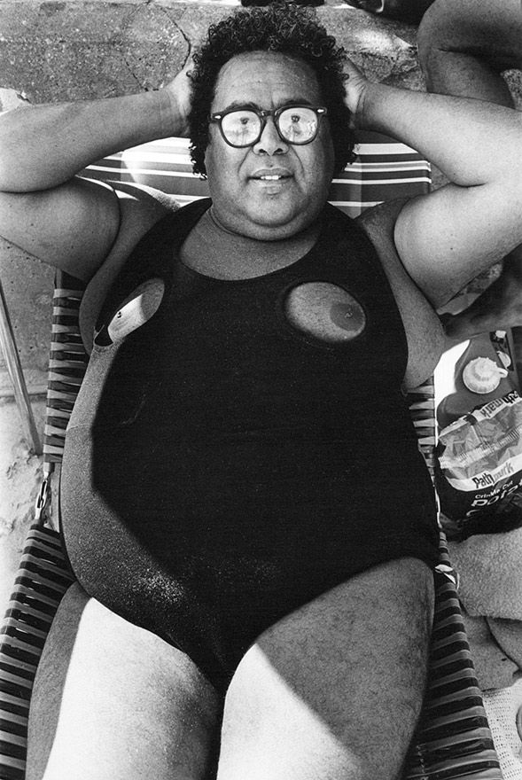 Man in a humorous swimsuit with eyes on chest, relaxes on a striped lounge chair at the beach. Smiling, hands behind head, snacks nearby.