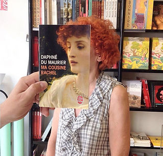 Hand holds book "DAPHNE DU MAURIER MA COUSINE RACHEL" over person's face.