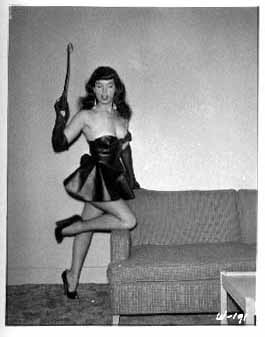 Bettie Page in black leather mini-dress, holding whip, leg on couch.