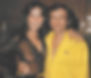 Linda Lovelace in a sheer black top and Hugh Hefner in a yellow shirt with a crest smile for the camera in a warmly lit wood-paneled room.