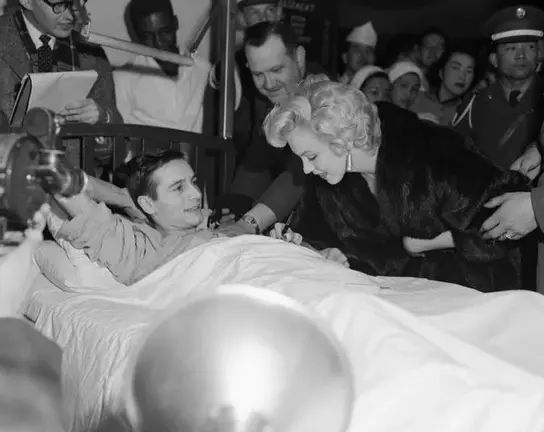 Blonde woman comforts man in hospital bed, onlookers observe.