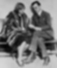A man and woman sit on a bench; the man reads a document. The woman wears a fur coat, looking pensive. Black and white vintage setting.