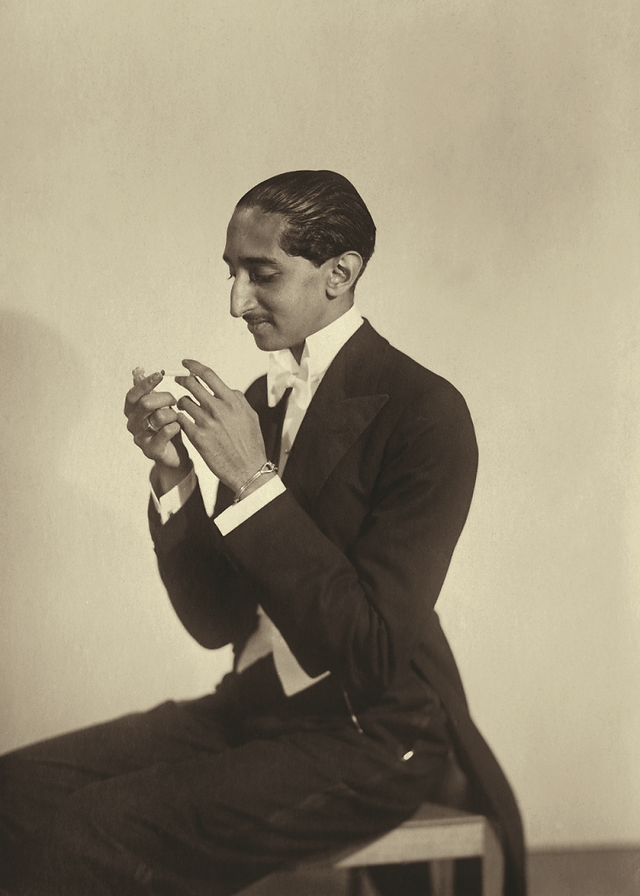 Man in tuxedo adjusting hands, looking down in sepia studio portrait.