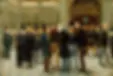Men in formal attire converse in a grand, ornate hall with a large clock and intricate wall designs. Some men wear top hats; mood is engaged.