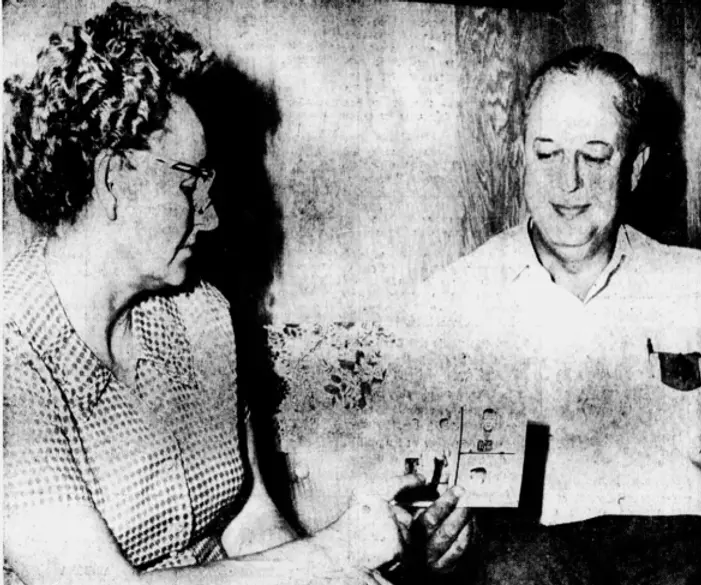 Grainy black-and-white photograph of an older couple seated together, with the woman holding and showing a small photograph to the man. The woman, wearing glasses and a patterned blouse, appears focused on the photo, while the man, dressed in a light shirt, smiles slightly as he looks at it. The setting has a homely, intimate feel, with subtle details in the background suggesting a casual, private moment.