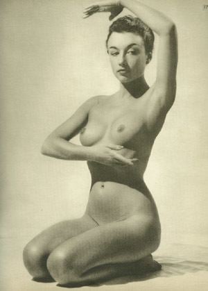 Nude woman delicately poses, one arm raised, hand covering breast.