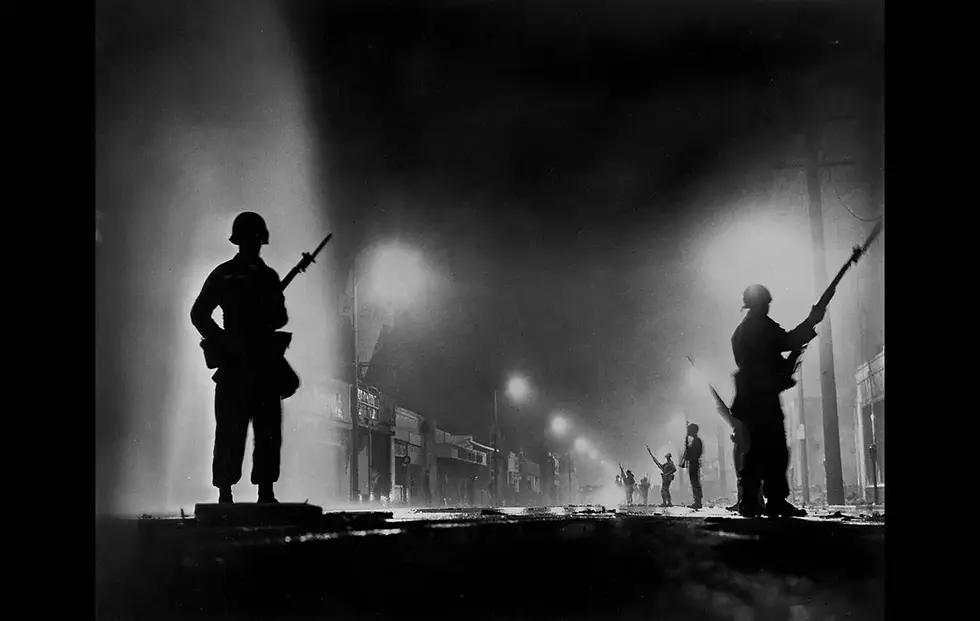 Silhouetted soldiers with rifles stand guard on a foggy street at night. Streetlights illuminate the background, creating a tense atmosphere.