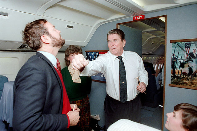 Ronald Reagan playfully punches bearded man near "EXIT" sign on plane.