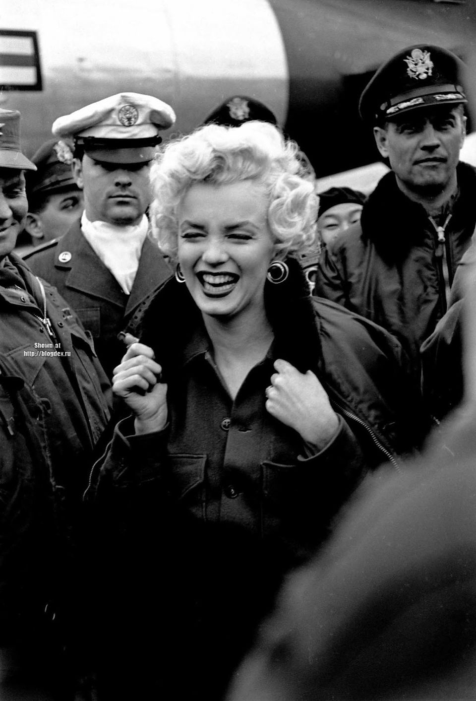 Marilyn Monroe laughing, surrounded by uniformed military personnel outdoors.