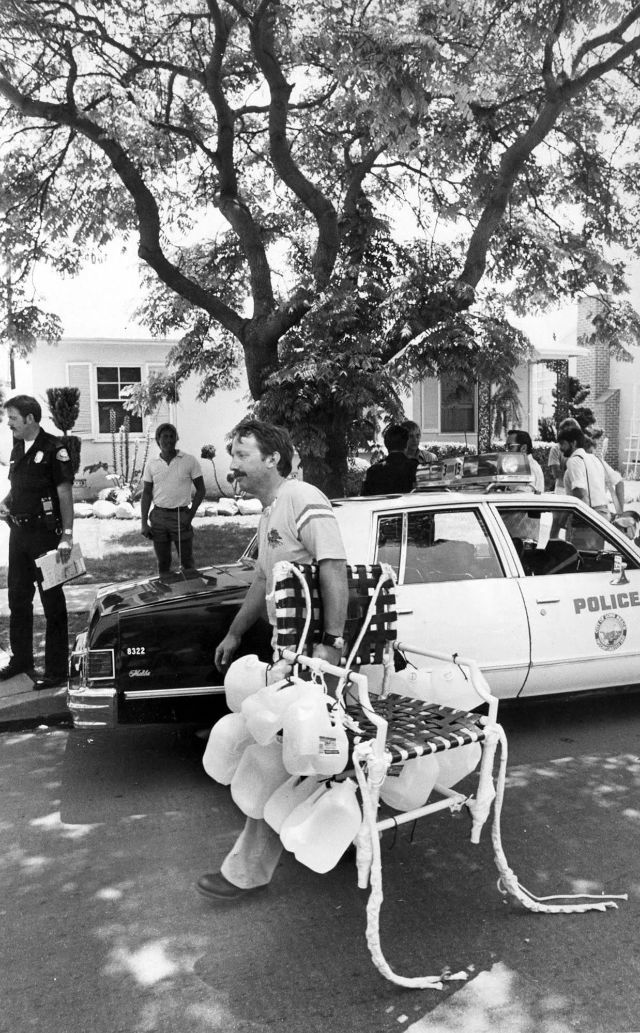 Lawnchair Larry carrying chair with jugs walks past police car under tree. Officers and onlookers nearby. Sunny day, suburban street scene.