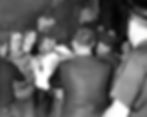 A black-and-white image shows a tense crowd scene with young people and police officers interacting. A brick building is visible in the background.