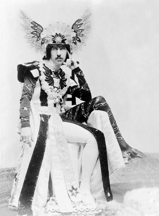 Person in ornate costume with winged headdress and intricate patterns sits on a chair in a vintage setting, exuding elegance and authority.