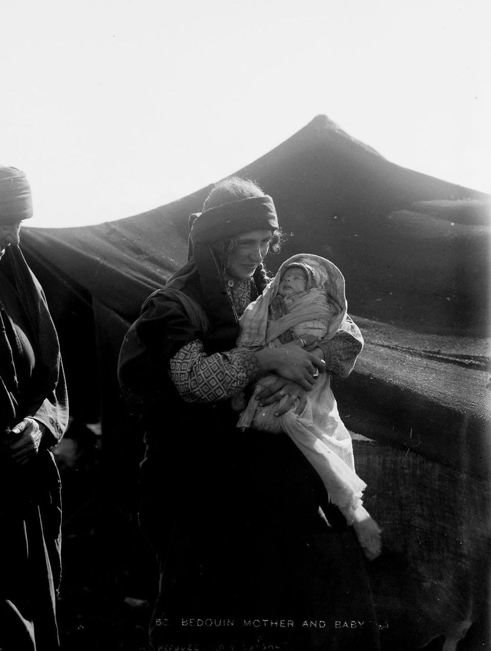 Black-and-white photograph of a Bedouin woman holding a swaddled infant in her arms, standing outside a large traditional tent. The woman wears patterned sleeves and a headscarf, gazing gently at the baby in soft morning or evening light. Taken in the early 20th century by the American Colony Photo Department.