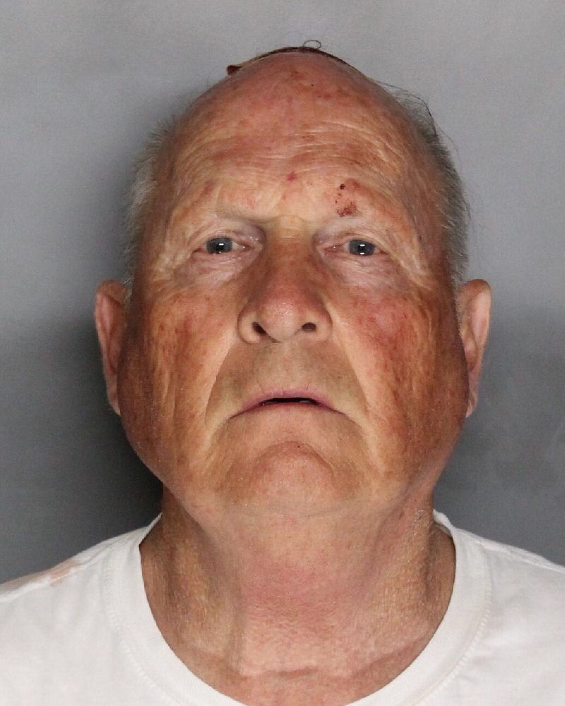 Elderly man with a serious expression against a gray background, wearing a white shirt. The image has a neutral, somber mood.