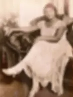 Woman seated on a plush chair in a lace dress, looking relaxed and poised. Vintage furniture in background, sepia-toned image.