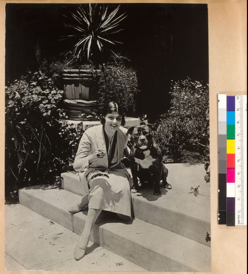Woman sitting on steps beside a dog, smiling with a cigarette in hand. Lush garden backdrop, large potted plant behind. Black and white.