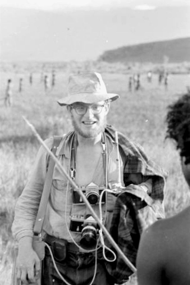 Man with cameras and hat smiles in a grassy field, holding a stick. Blurry background with figures scattered in the distance. Black and white.
