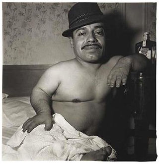 Man with mustache and hat, shirtless, leaning on chair, draped in sheet.