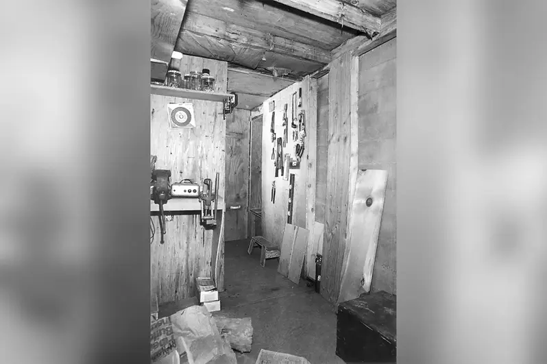 A dimly lit workshop with wooden walls; tools hang neatly, a drill press sits on a bench. Boxes and boards scatter the floor. Mood: rustic.