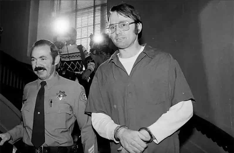 Officer escorts a handcuffed man down a dim hallway. Reporters with flash cameras are present. The mood is serious, and it's a black-and-white photo.