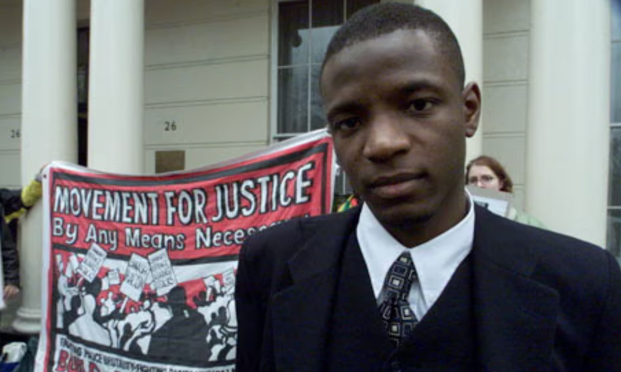 Duwayne Brooks in a suit stands in front of a protest banner reading "Movement for Justice By Any Means Necessary" with a serious expression.