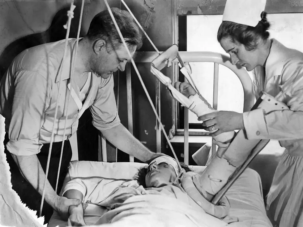 A nurse and a man assist a patient with an arm cast in a hospital bed using traction equipment, conveying a serious mood. Black and white.