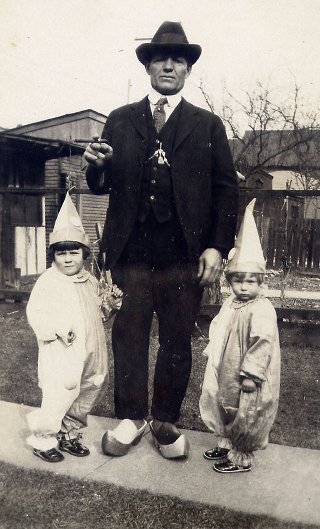 Man in suit and hat stands with two children in clown costumes. Wooden clogs, backyard setting, bare trees. Vintage, playful mood.