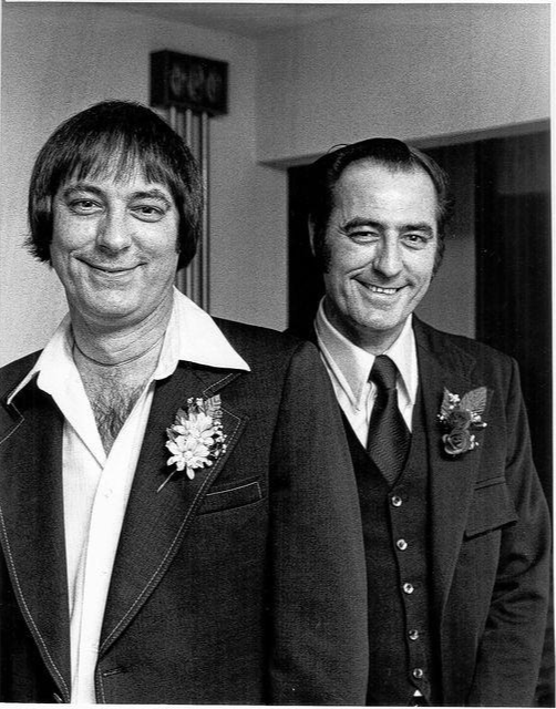The Jim Twins smiling men in suits with floral boutonnieres stand indoors, one slightly in front of the other. The mood is cheerful and formal.