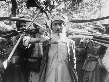 A man with a beard stands among armed men in turbans brandishing rifles and swords. They appear defiant in a sunlit wooded area.