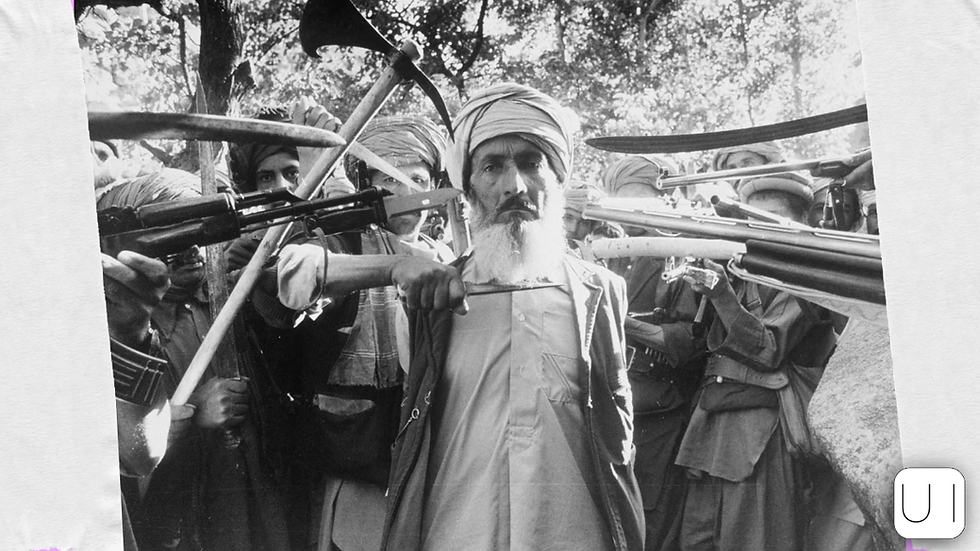 A man with a beard stands among armed men in turbans brandishing rifles and swords. They appear defiant in a sunlit wooded area.