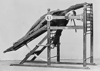 Man in suit lies face down on wooden inversion apparatus, K1 visible.