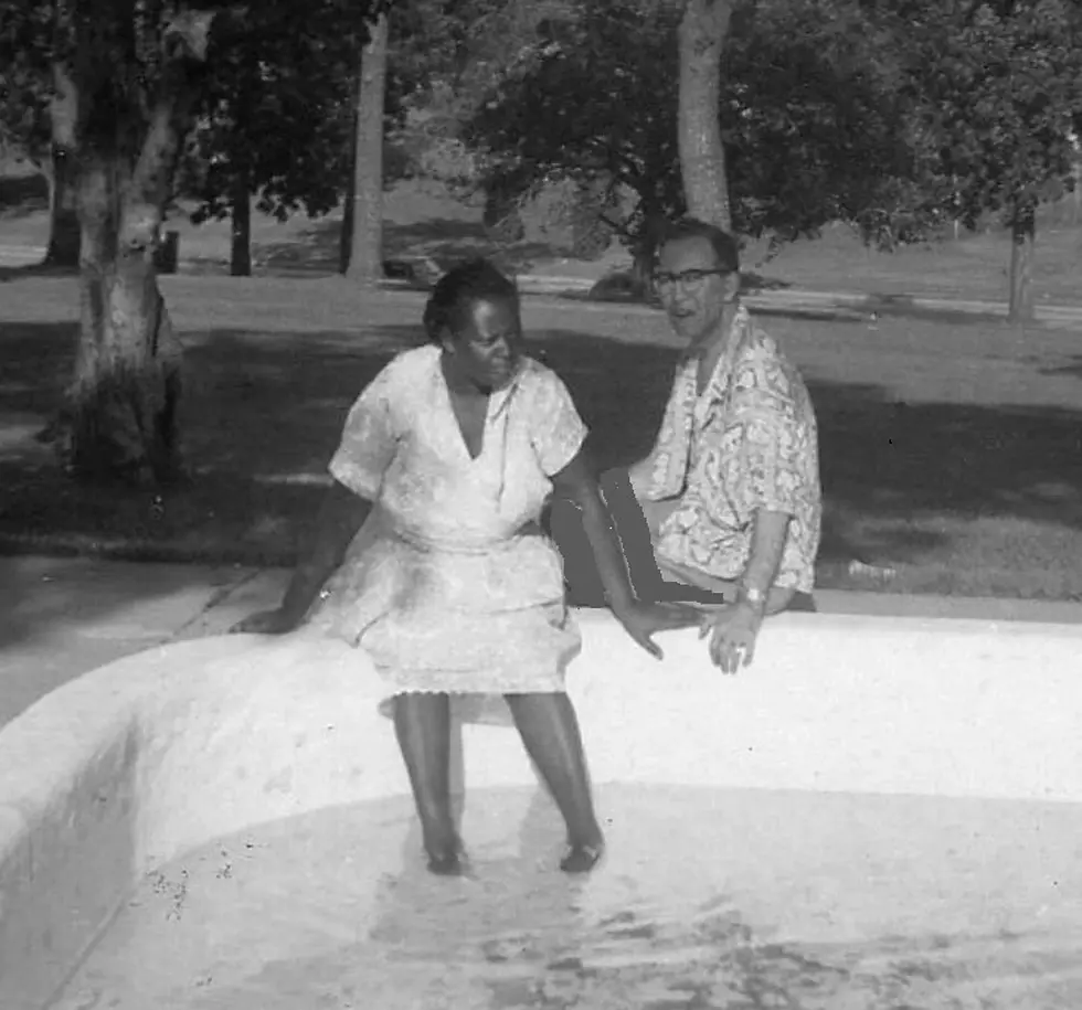 Two people sit on the edge of an empty pool, laughing. The scene is in a park with trees in the background. The mood is relaxed.