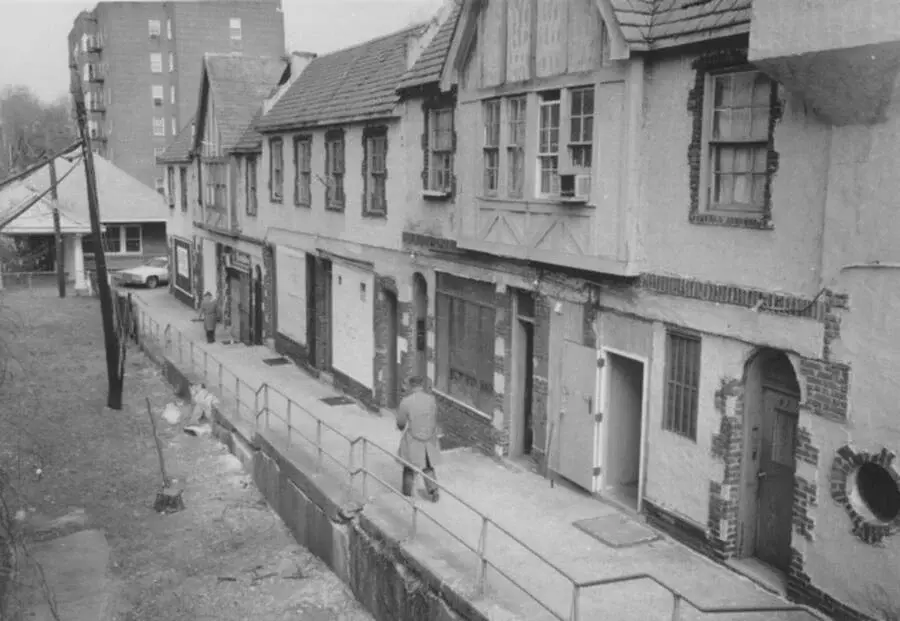 Black and white image of a person walking along a narrow alley beside old brick buildings. Another person is near a distant car. Urban setting.
