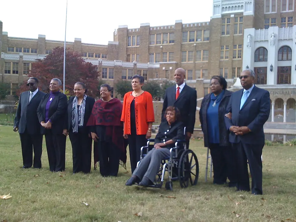 Group of nine people pose in front of a historic building. One person is seated in a wheelchair. The mood is solemn, with autumn foliage.